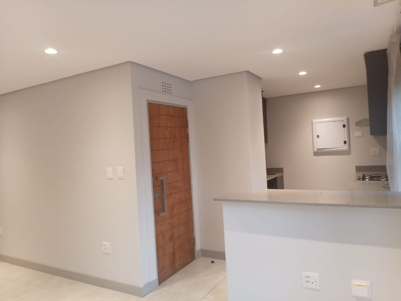 Modern open-plan apartment interior featuring a neutral color palette, recessed ceiling lighting, a wooden entrance door, and a functional kitchen breakfast bar.