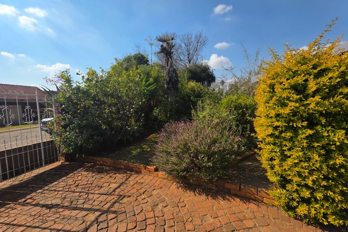 A front garden area featuring established shrubs, trees, and a brick-paved driveway section enclosed by a metal gate.