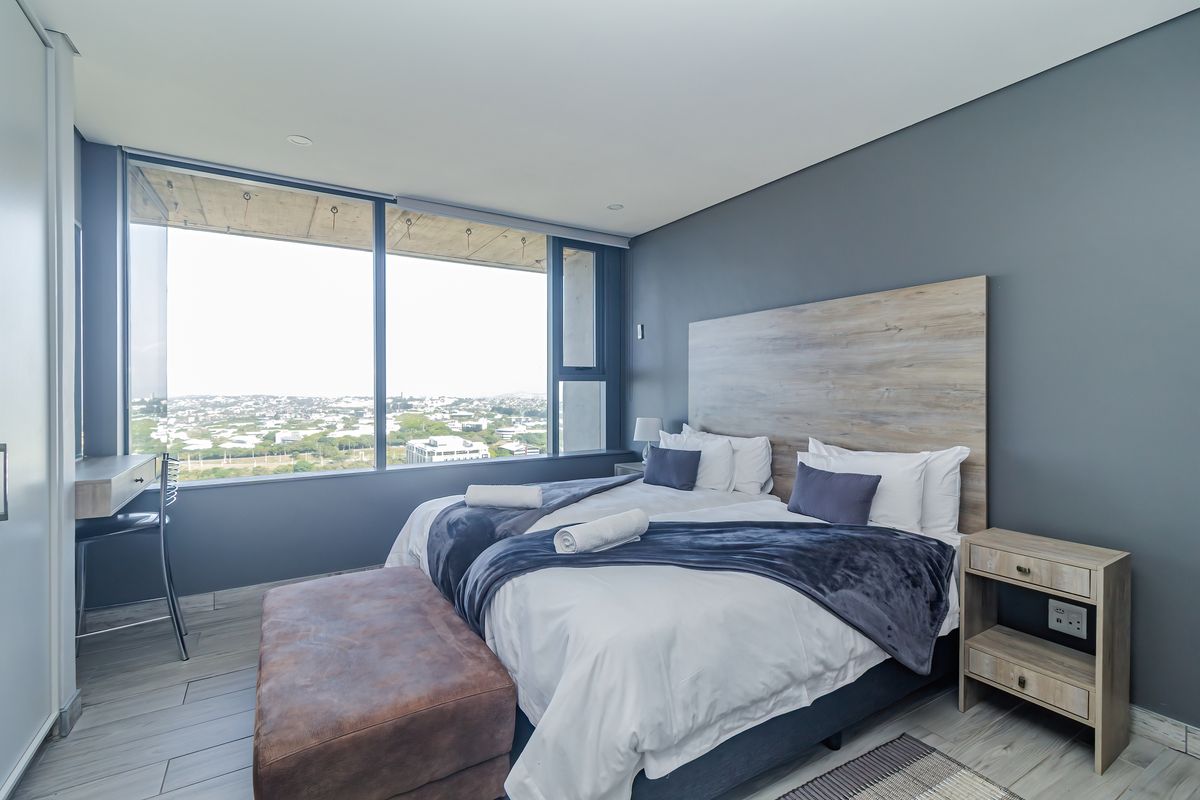 Modern bedroom featuring large floor-to-ceiling windows with expansive city views, neutral grey walls, and contemporary tiled flooring.