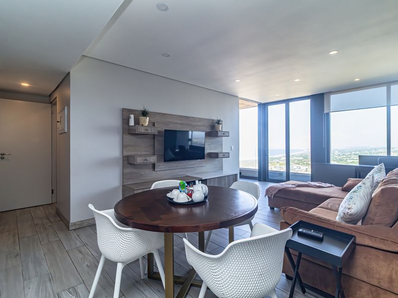 Modern open-plan living area featuring wood-look tile flooring, a built-in media console, and expansive floor-to-ceiling windows with panoramic coastal views.