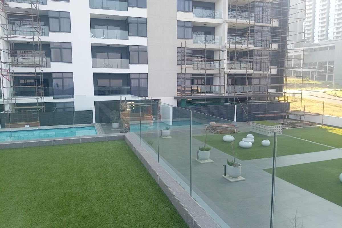 View of the apartment complex exterior featuring a swimming pool, landscaped lawn area, and glass balustrades, currently undergoing building maintenance and construction.