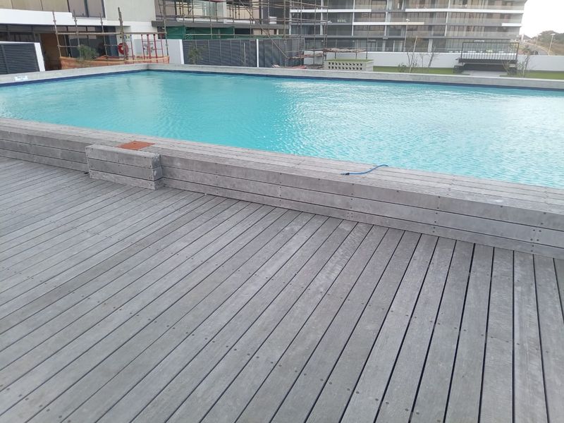 A communal swimming pool area featuring a large rectangular pool with timber decking surround, set within a modern apartment complex.