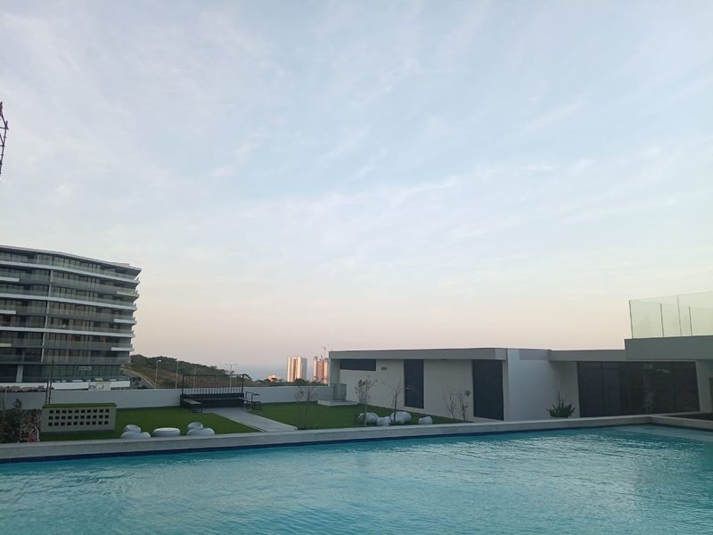 A rooftop swimming pool area featuring a clear blue pool, landscaped lawn, and modern building architecture with expansive city and horizon views.