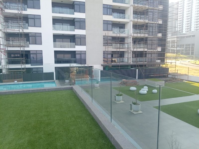 View of the apartment complex exterior featuring a swimming pool, landscaped lawn area, and glass balustrades, currently undergoing building maintenance and construction.