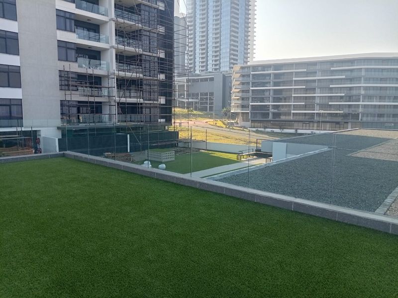 A view of a landscaped rooftop terrace featuring artificial turf and glass balustrades, overlooking surrounding modern apartment buildings.