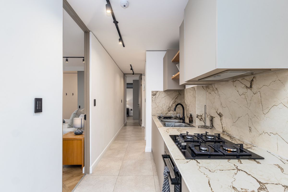 Modern galley-style kitchen featuring stone countertops, marble-look splashback, gas hob, and integrated cabinetry, leading into a well-lit hallway.