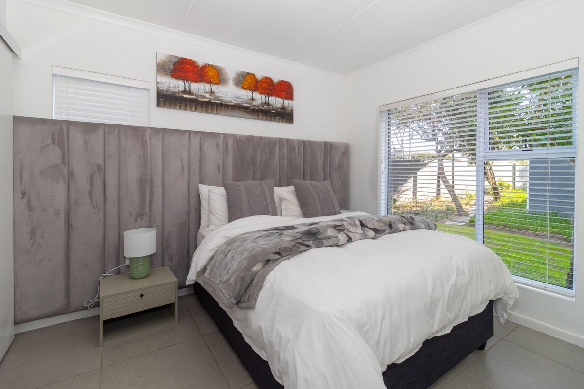 A bright bedroom featuring a large upholstered headboard, tiled flooring, and large windows with fitted blinds overlooking the garden.