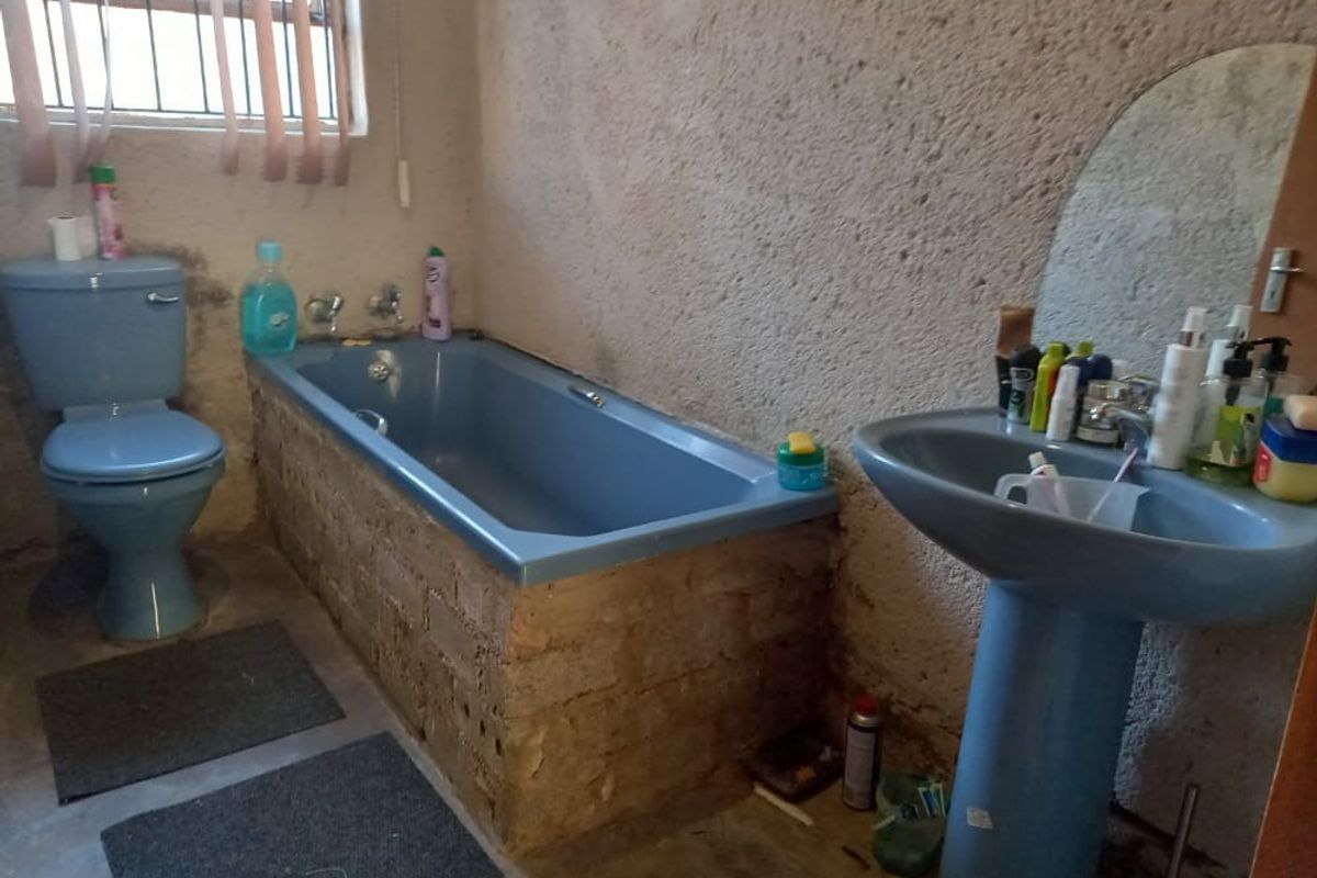 A dated bathroom featuring blue sanitary ware, including a pedestal basin, toilet, and bathtub with an unfinished brick surround and rough plaster walls.