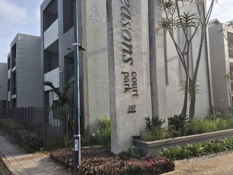 Street-level view of the Seasons Court apartment complex entrance, featuring a prominent concrete signage pillar and landscaped garden beds.