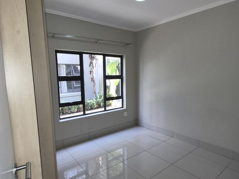 A bright, empty bedroom featuring neutral tiled flooring, a large window with aluminium frames, and a ceiling-mounted light fixture.