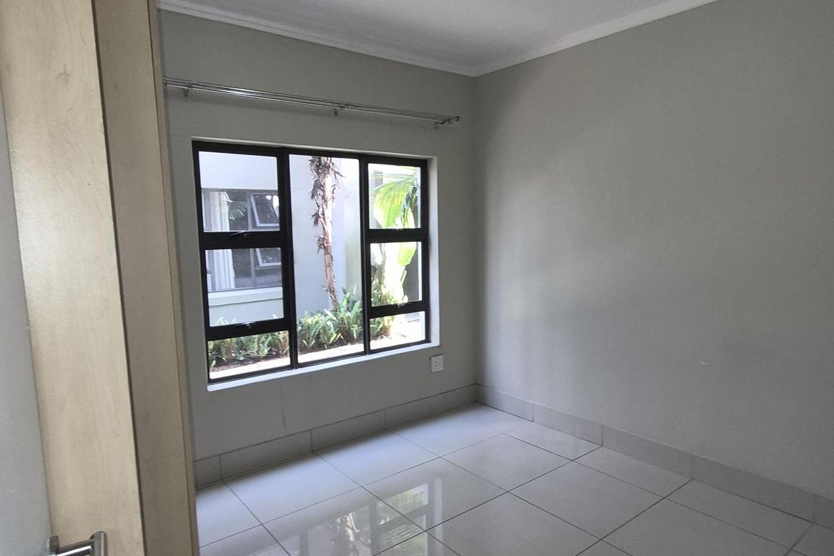 A bright, empty bedroom featuring neutral tiled flooring, a large window with aluminium frames, and a ceiling-mounted light fixture.