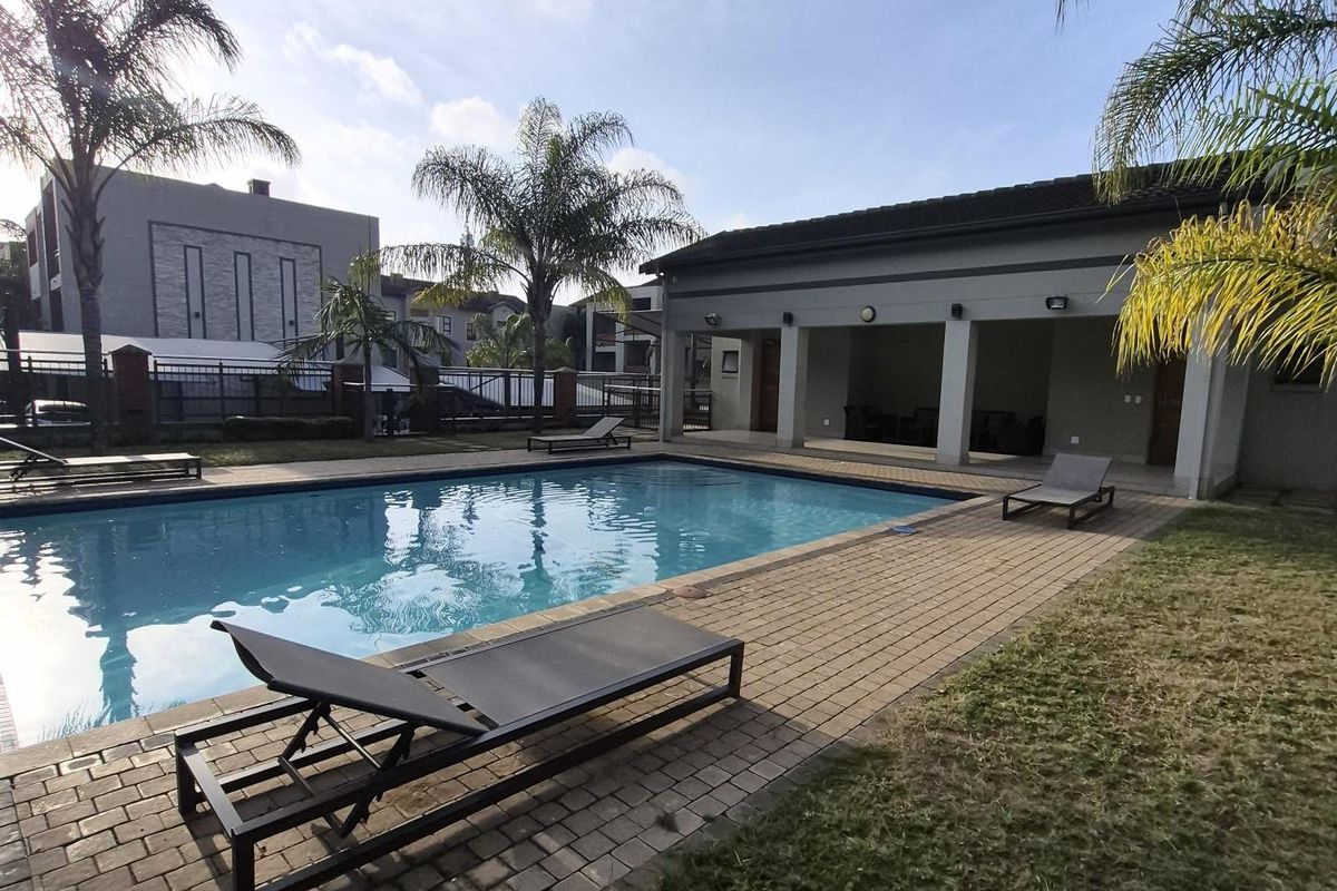 A well-maintained communal swimming pool area featuring paved decking, a covered patio space, and landscaped surroundings within the apartment complex.