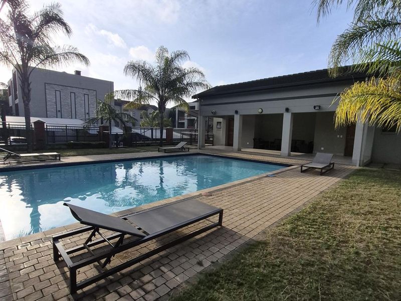 A well-maintained communal swimming pool area featuring paved decking, a covered patio space, and landscaped surroundings within the apartment complex.