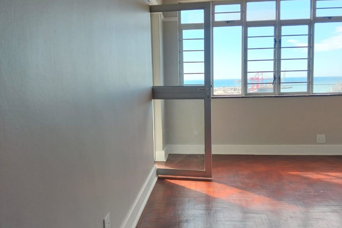An empty living area featuring classic parquet flooring, neutral walls, and large windows offering a scenic coastal view.
