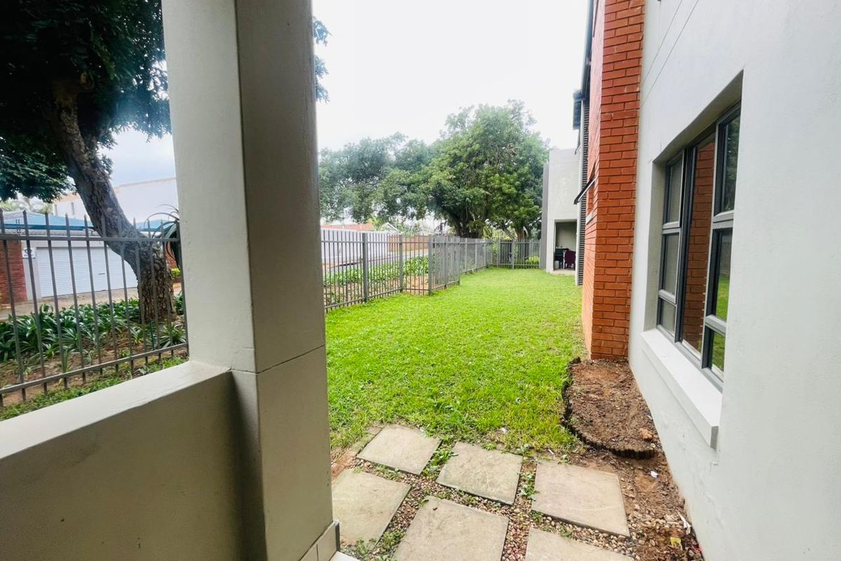 A private rear garden area featuring a small lawn, paved walkway, and perimeter fencing, accessible from the side of the apartment building.