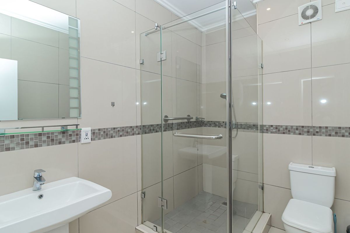 A clean, modern bathroom featuring a glass-enclosed shower, pedestal washbasin, and neutral ceramic wall tiles with a decorative mosaic border.