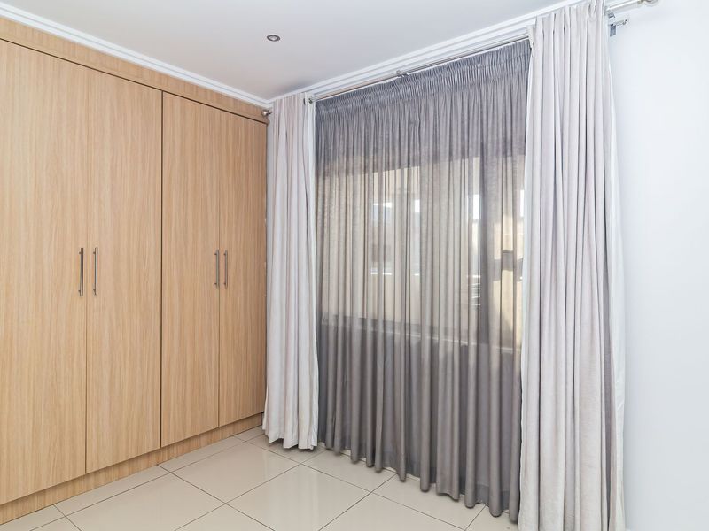 A clean, bright bedroom featuring light wood-finish built-in wardrobes, neutral tiled flooring, and modern recessed lighting.