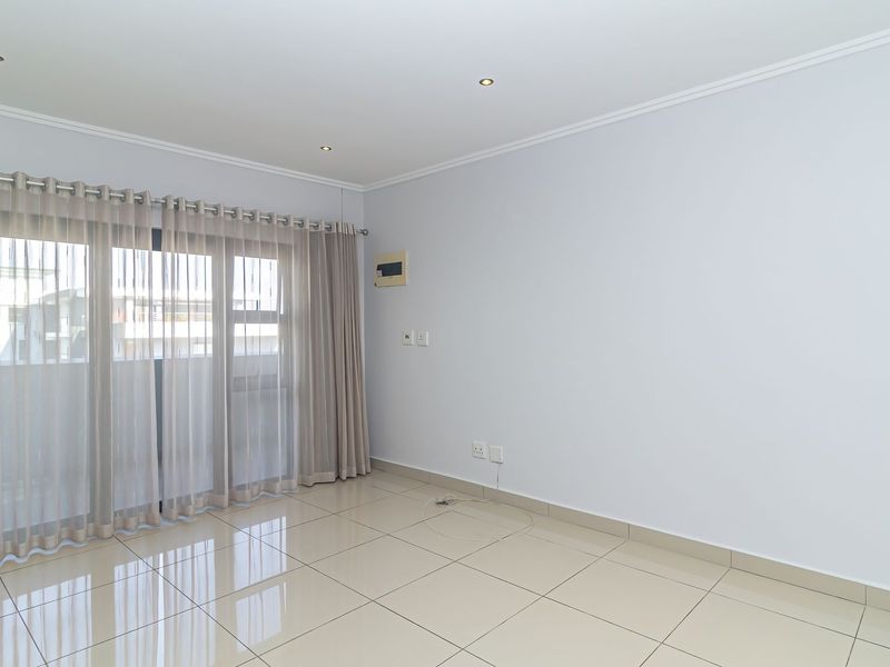 A bright, empty living room featuring neutral tiled flooring, recessed downlights, and a large sliding glass door providing access to the balcony.