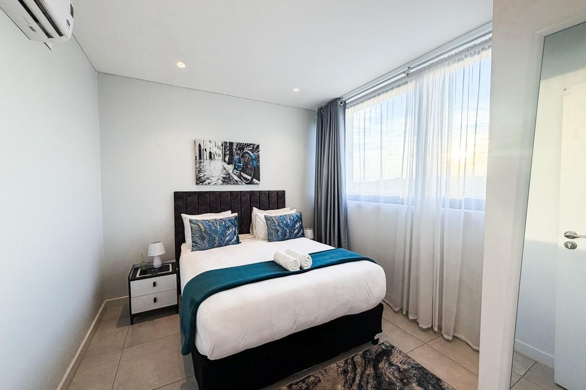 A modern bedroom featuring tiled flooring, recessed ceiling lighting, and a wall-mounted air conditioning unit, with large windows providing natural light.