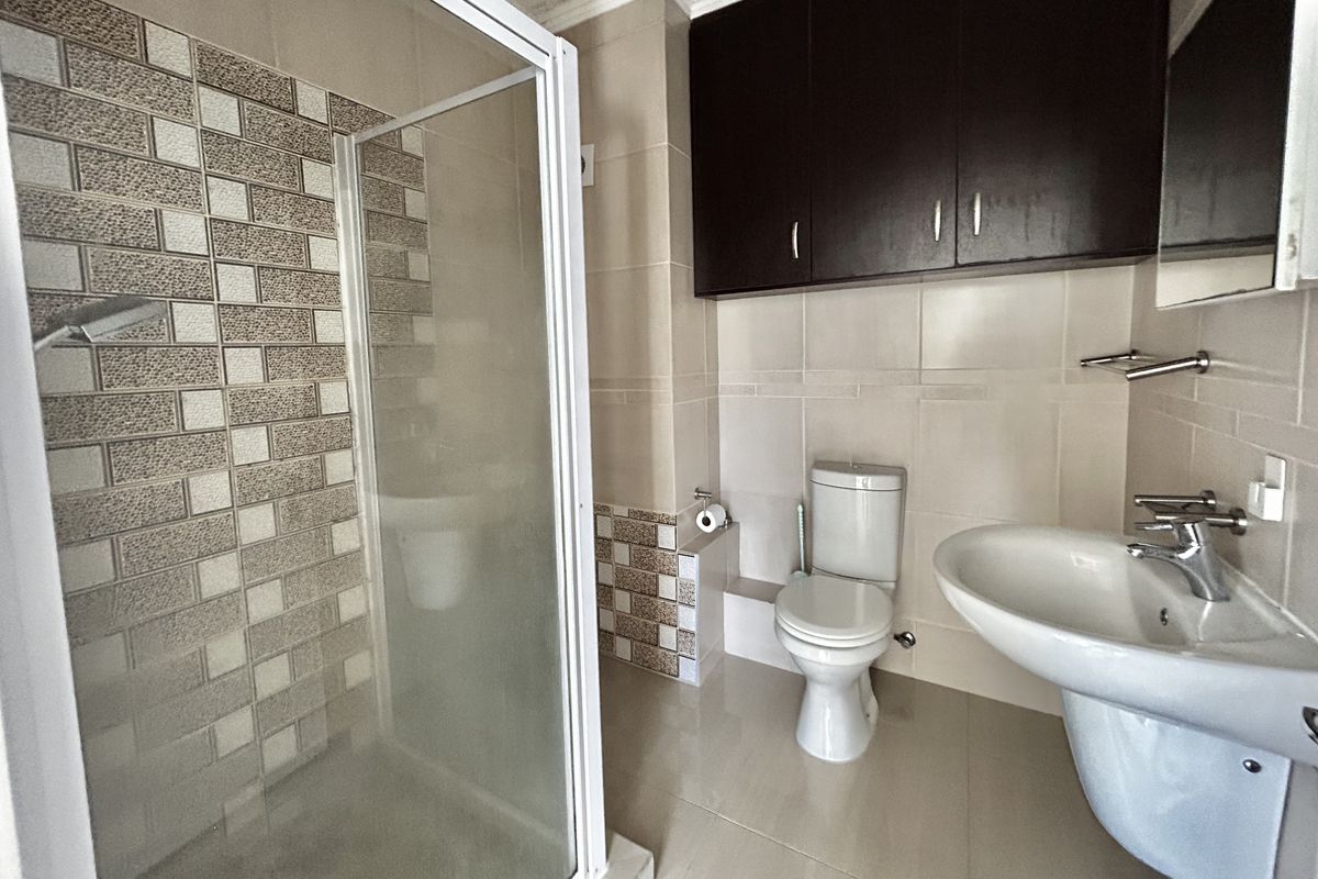 A functional bathroom featuring a glass-enclosed shower, pedestal basin, toilet, and dark wood-finish wall-mounted cabinetry with tiled walls and flooring.