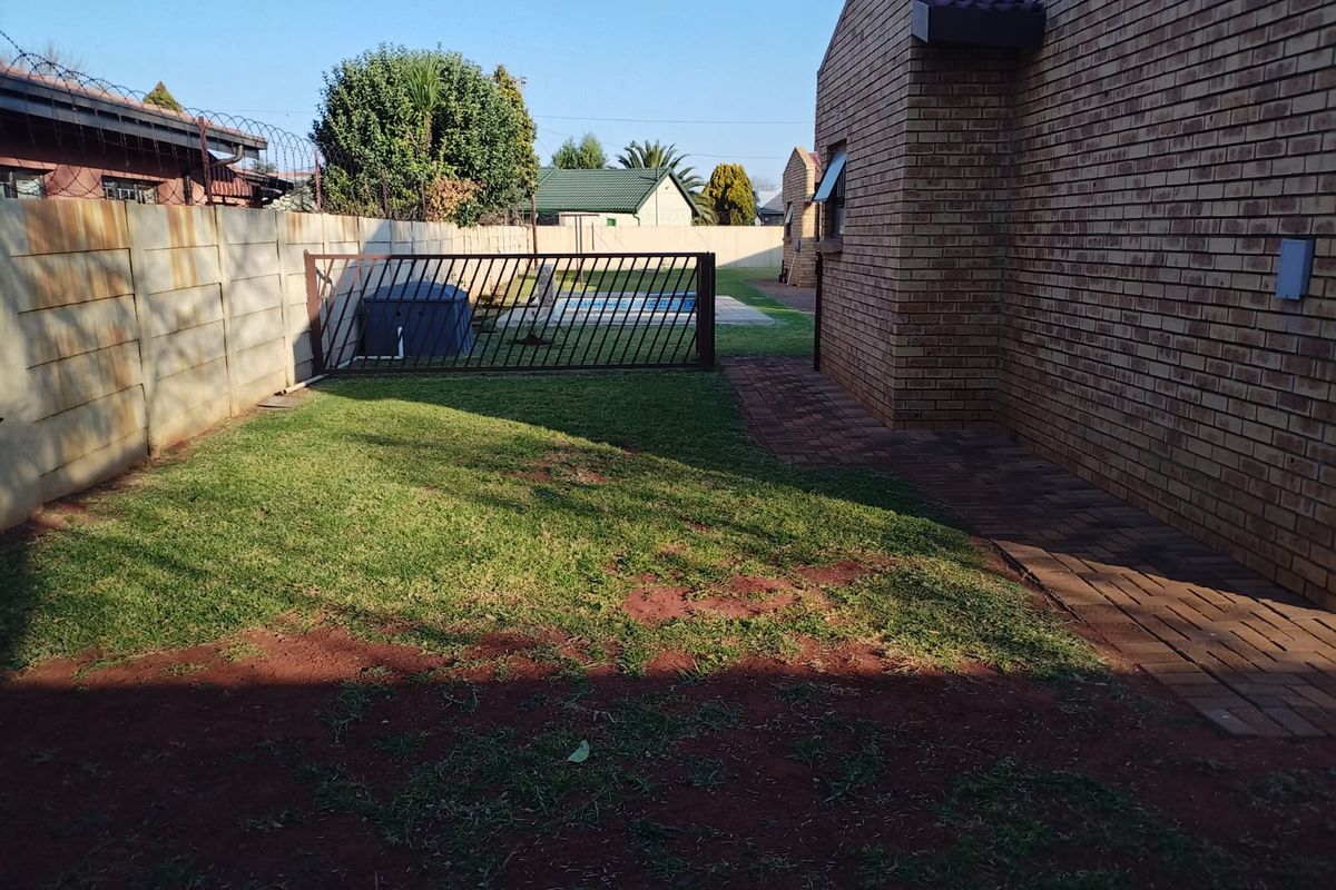 A view of the rear garden area featuring a brick exterior, paved walkway, and a metal security gate leading to the pool area.