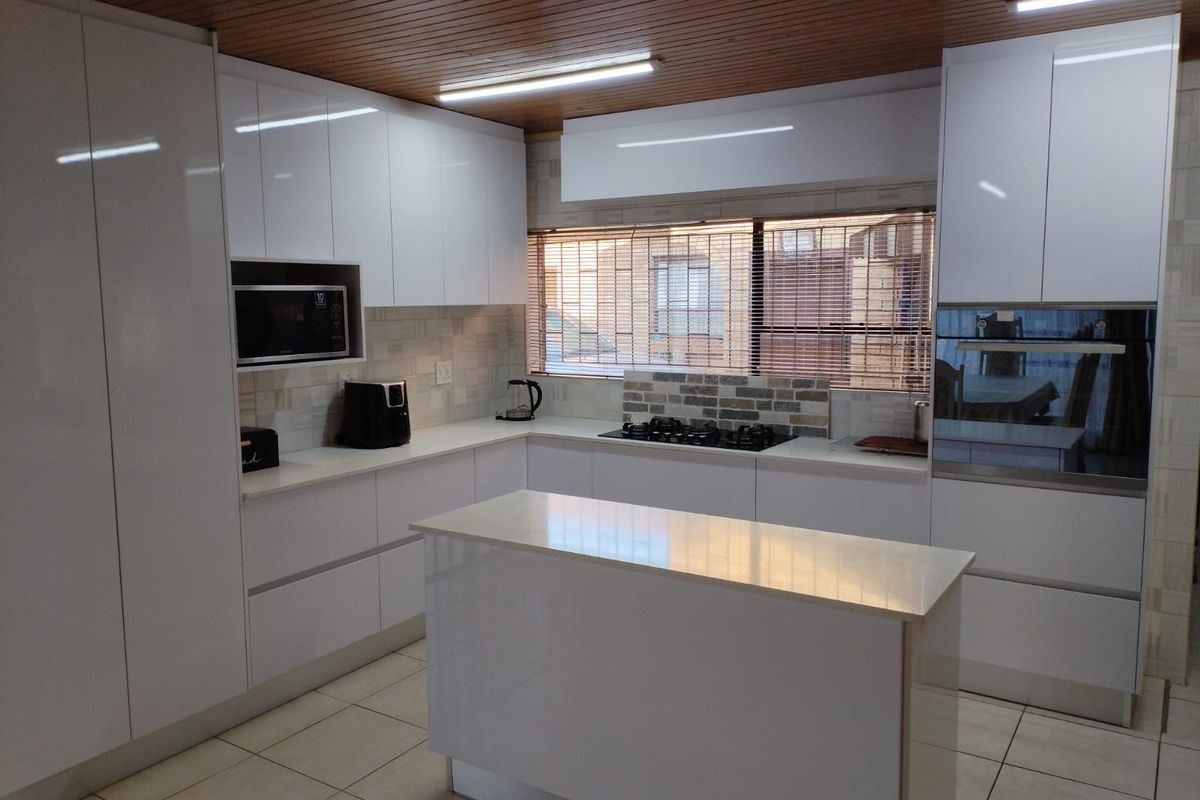 Modern kitchen featuring high-gloss white cabinetry, a central island, built-in oven, gas hob, and tiled flooring under a timber-paneled ceiling.
