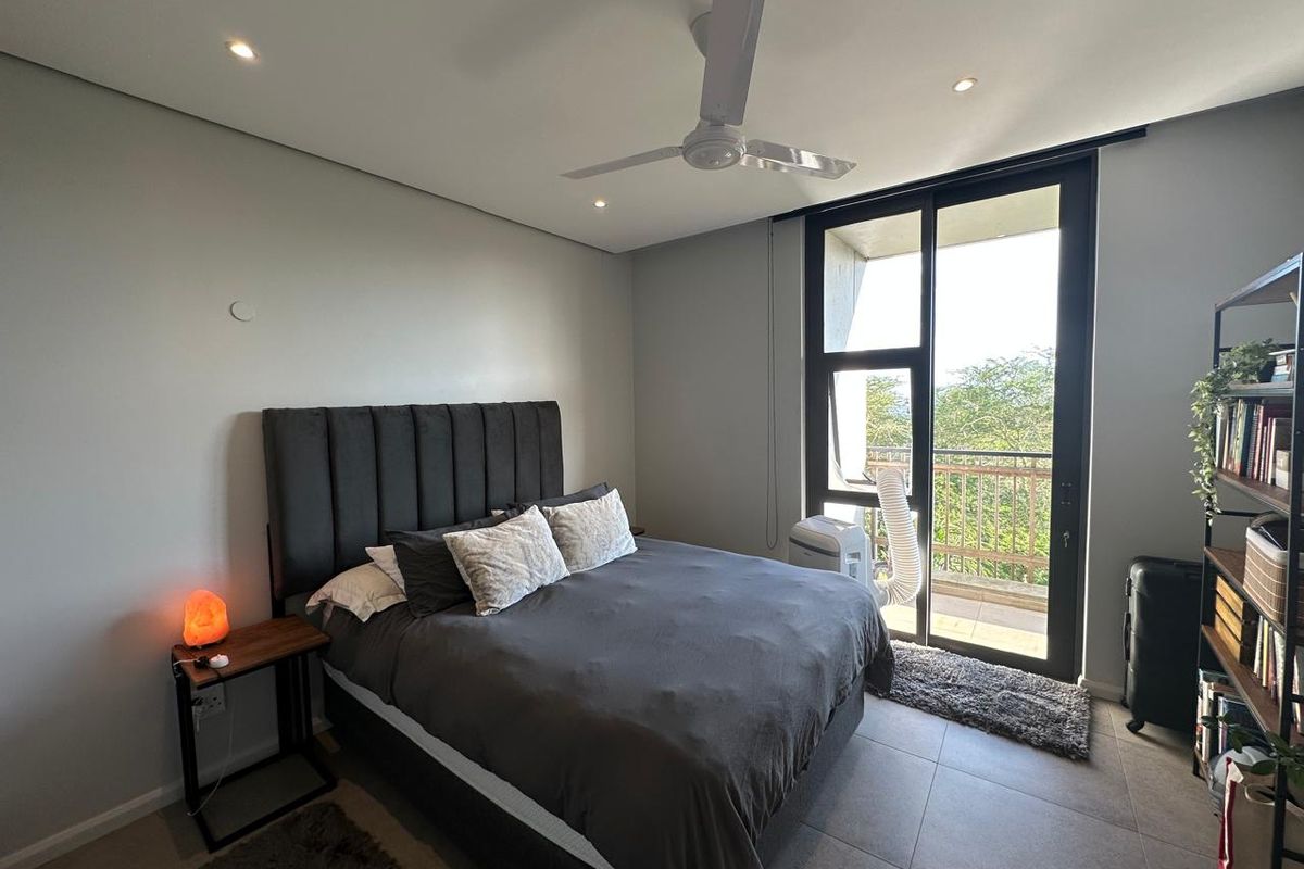 A modern bedroom featuring tiled flooring, a ceiling fan, and large sliding glass doors that open onto a private balcony with greenery views.
