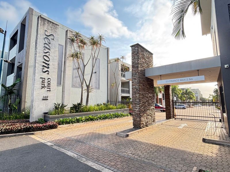 The main entrance to Seasons Court, featuring a paved driveway, stone-clad gate pillar, security boom gate, and landscaped garden beds.