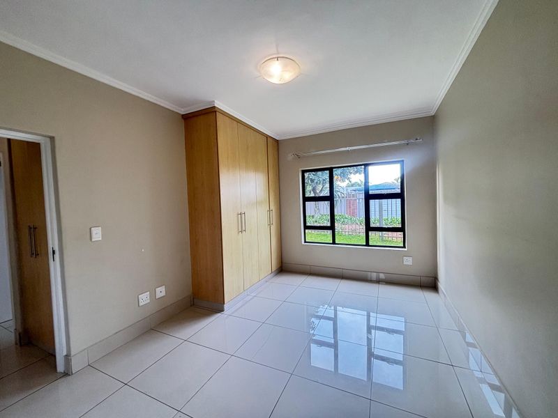 A bright, empty bedroom featuring neutral tiled flooring, built-in wooden cupboards, and a large window providing natural light.
