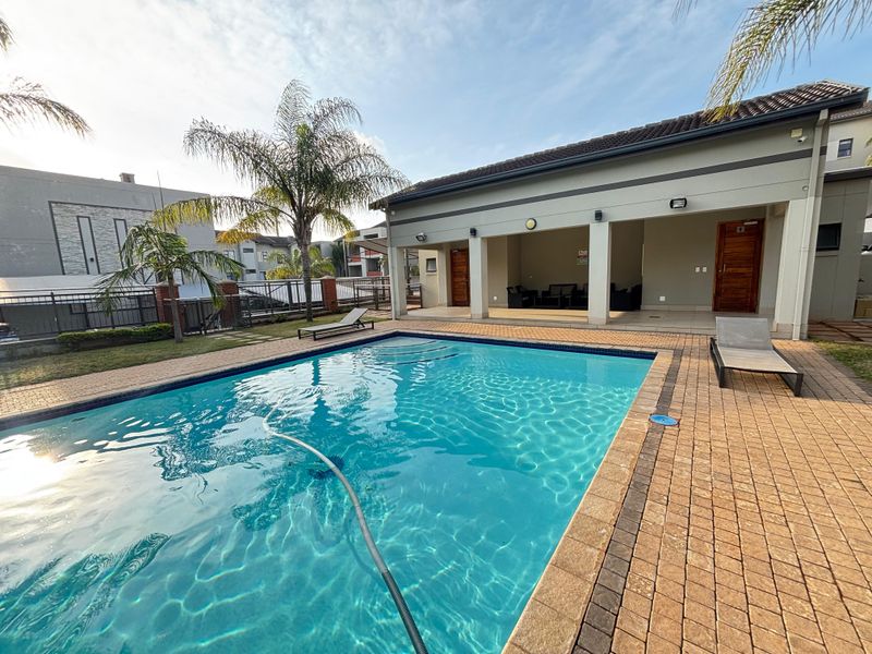 A well-maintained communal pool area featuring a paved deck, covered patio space, and landscaped surroundings within the apartment complex.