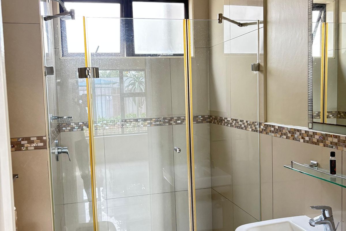 A functional bathroom featuring a glass-enclosed shower, pedestal basin, and neutral wall tiling with a decorative mosaic border.