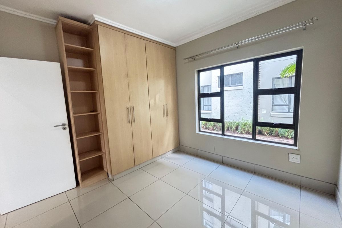 A bright bedroom featuring large format floor tiles, built-in wooden cabinetry with integrated shelving, and a window with a view of the exterior.