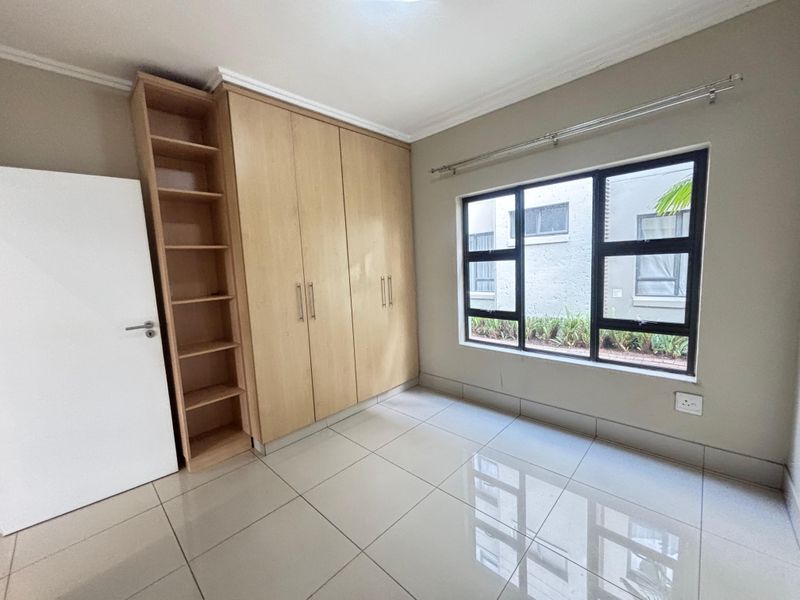A bright bedroom featuring large format floor tiles, built-in wooden cabinetry with integrated shelving, and a window with a view of the exterior.