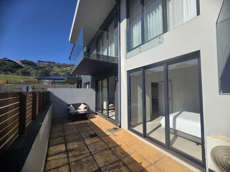 A modern ground floor patio area featuring tiled flooring, glass balustrades, and large sliding doors providing seamless access to the interior living space.