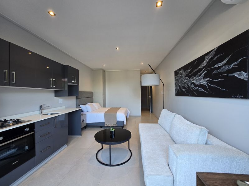A modern open-plan studio apartment featuring a sleek grey kitchen with built-in appliances, tiled flooring, and integrated downlighting throughout the living and sleeping area.