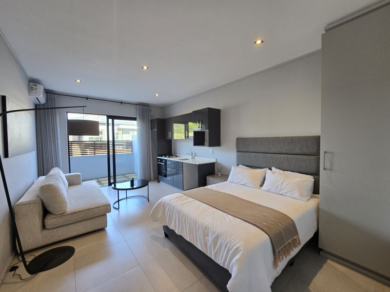 A modern open-plan studio apartment featuring a compact kitchenette, tiled flooring, recessed lighting, and sliding glass doors leading to a private balcony.