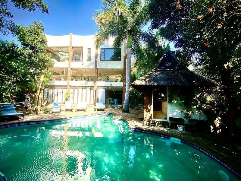 A multi-level rear exterior view featuring a swimming pool, landscaped garden, and a traditional thatched lapa entertainment area.