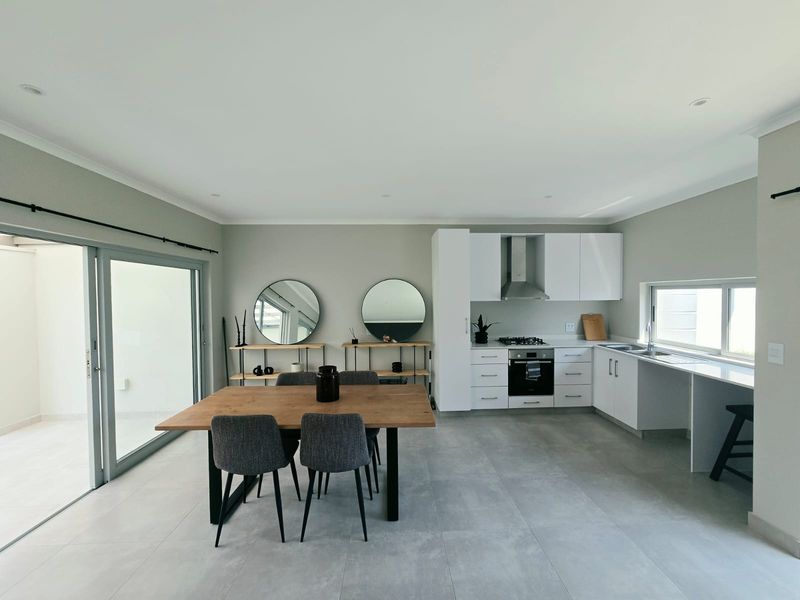 Modern open-plan kitchen and dining area featuring sleek white cabinetry, integrated appliances, large format floor tiles, and seamless access to an outdoor patio.