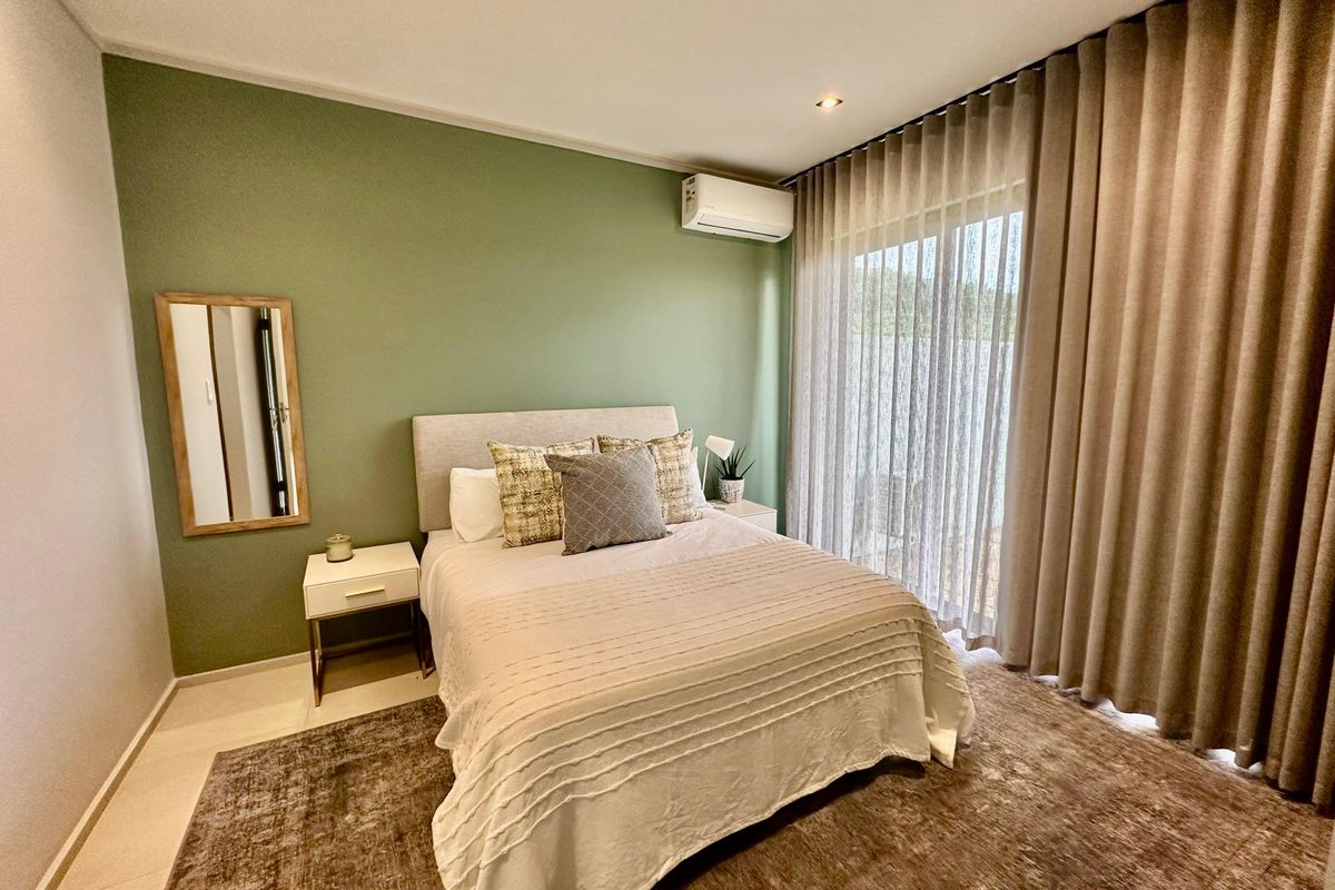 A well-maintained bedroom featuring a soft green accent wall, split-system air conditioning unit, and floor-to-ceiling curtains leading to an outdoor area.