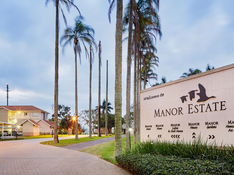Entrance view of Manor Estate featuring a paved driveway, landscaped palm trees, and estate signage at dusk.