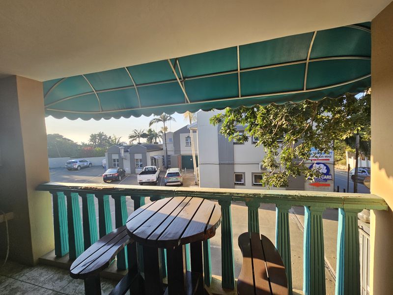 A covered balcony featuring a green awning and a decorative balustrade, overlooking the apartment complex parking area.