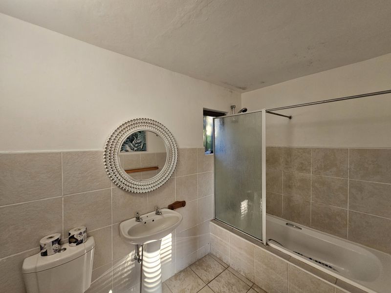 A functional bathroom featuring a pedestal sink, a bathtub with a glass shower screen, and neutral wall and floor tiling.