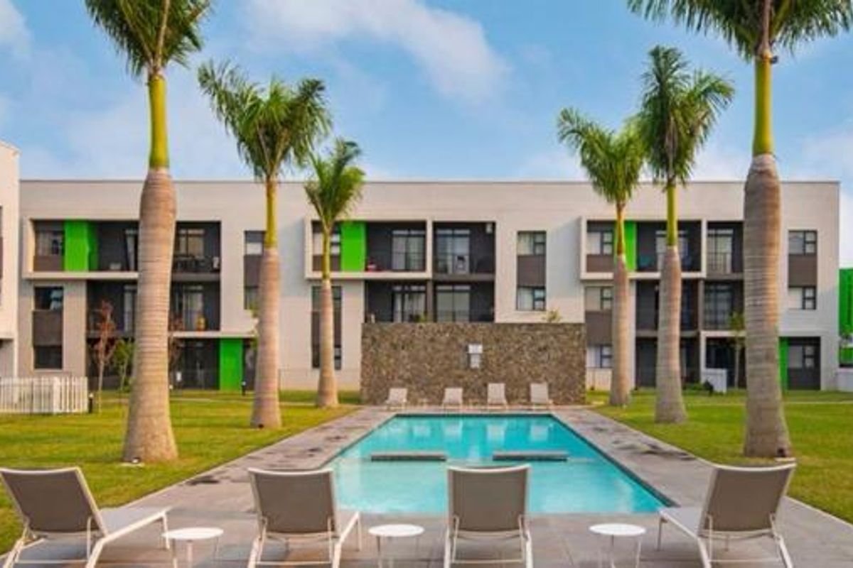 A well-maintained communal swimming pool area surrounded by landscaped lawns and palm trees, set against the modern facade of an apartment complex.