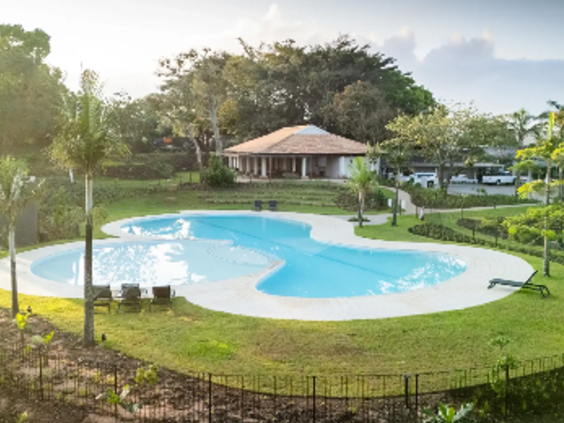 An elevated view of a well-maintained swimming pool set within a landscaped garden area, featuring paved surrounds and perimeter fencing.