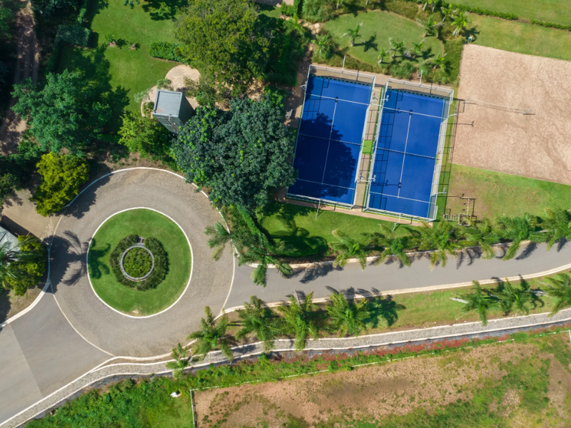 Aerial view of a residential property featuring two blue tennis courts, a sand volleyball court, a paved roundabout, and well-maintained landscaped grounds.