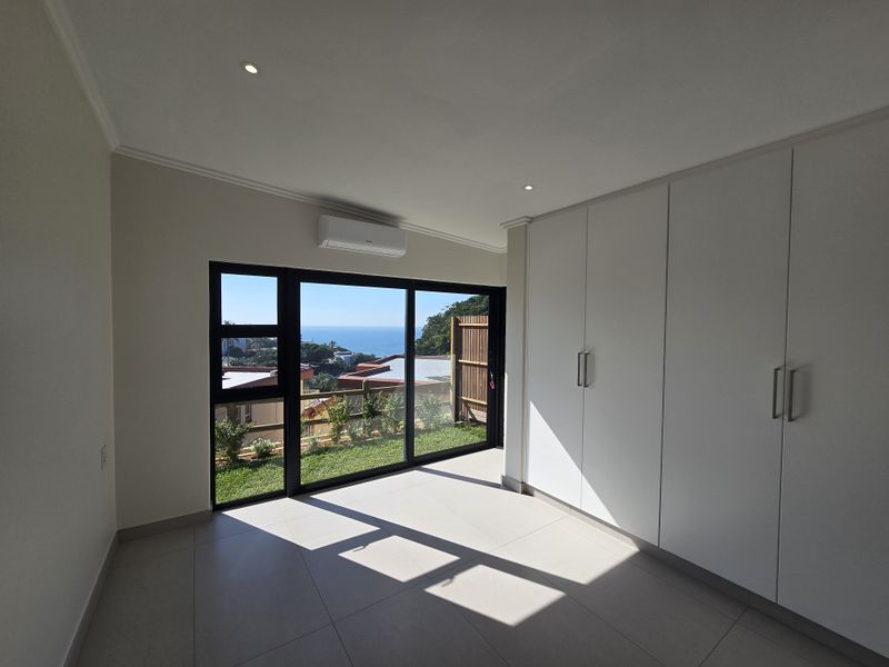 Bright, modern bedroom featuring built-in cupboards, tiled flooring, and a large sliding glass door opening to a garden with ocean views.