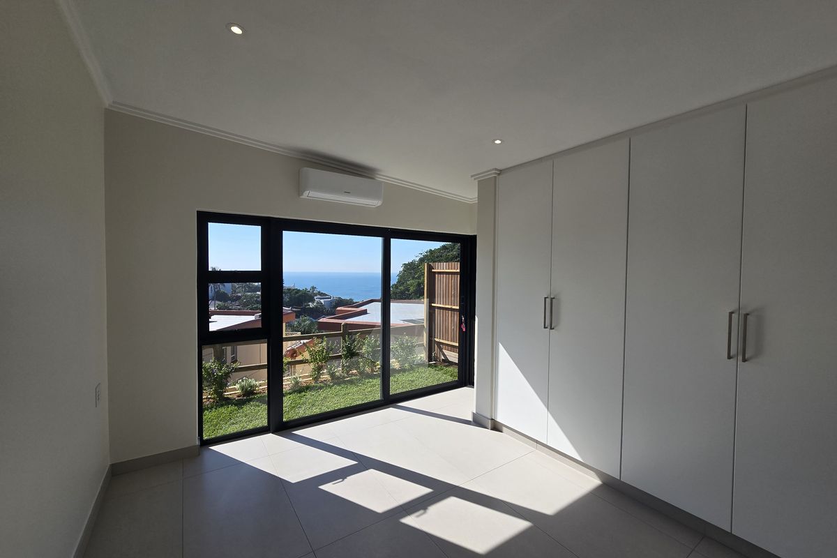 Bright, modern bedroom featuring built-in cupboards, tiled flooring, and a large sliding glass door opening to a garden with ocean views.