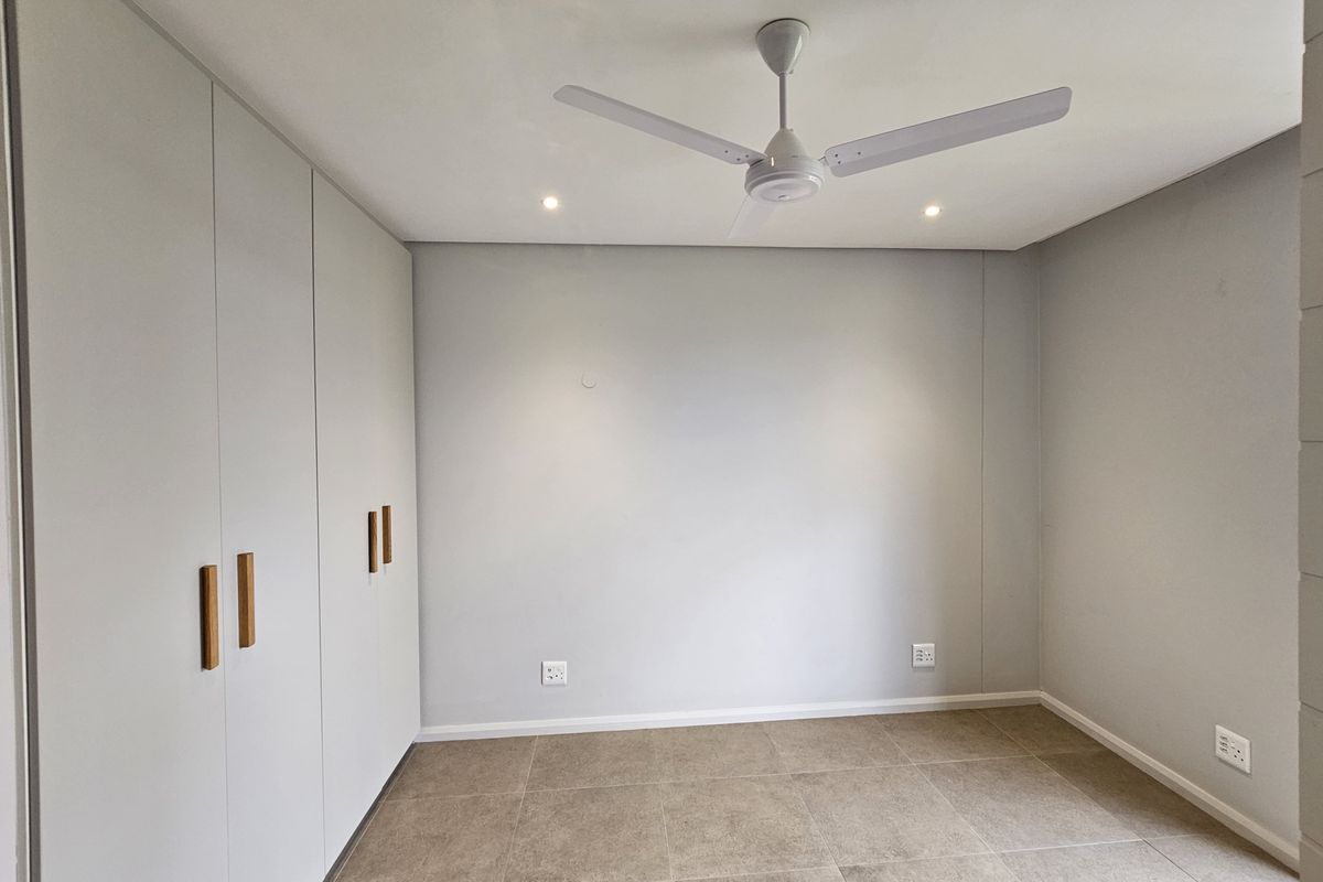 A clean, modern bedroom featuring neutral tiled flooring, built-in cupboards with wooden handles, a ceiling fan, and recessed lighting.