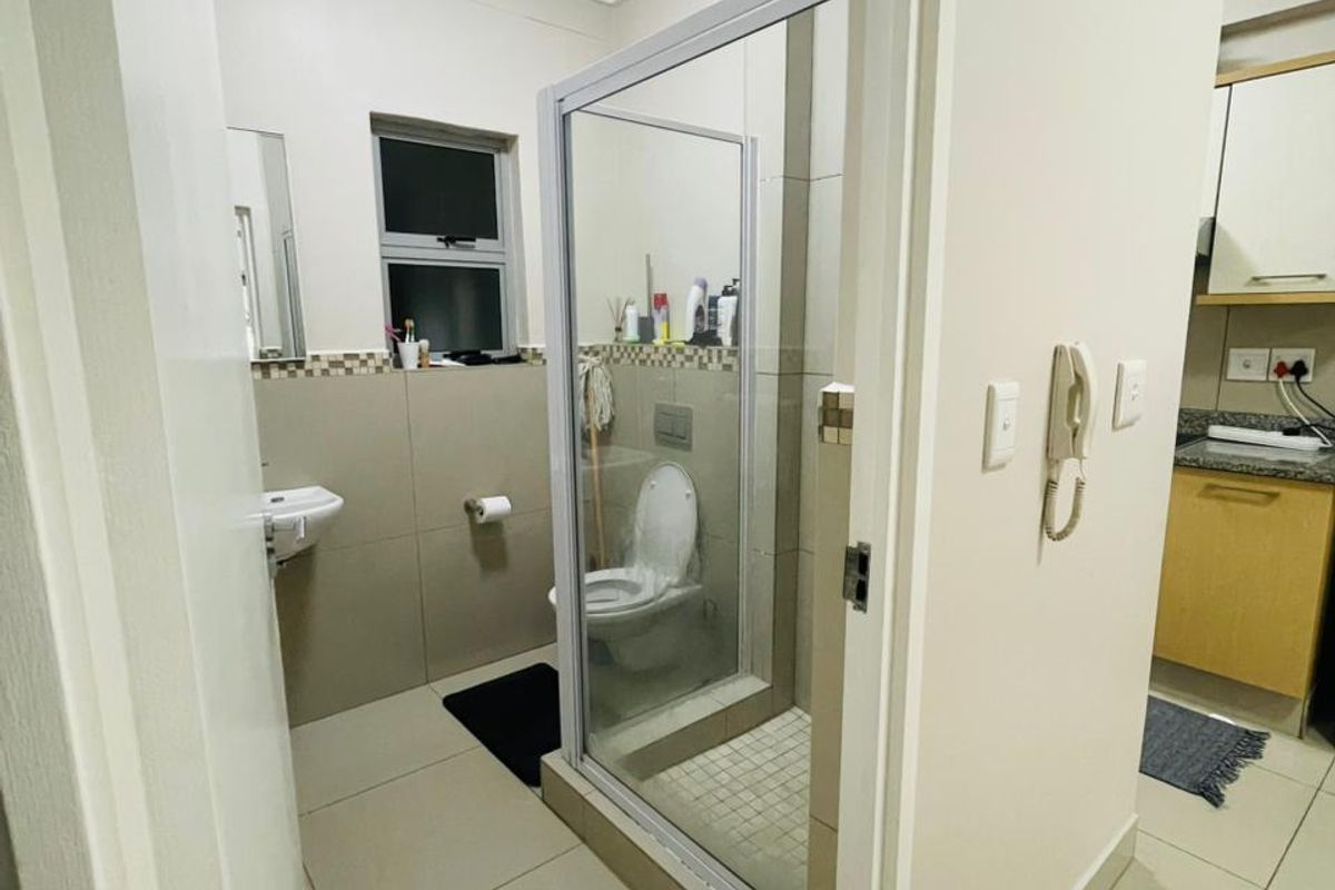 A functional bathroom featuring a glass-enclosed shower, wall-mounted basin, and neutral tiled finishes, with an adjacent view into the kitchen area.