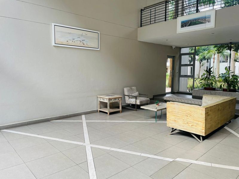Spacious double-volume living area featuring large format floor tiles, a mezzanine level with metal balustrade, and abundant natural light through expansive windows.
